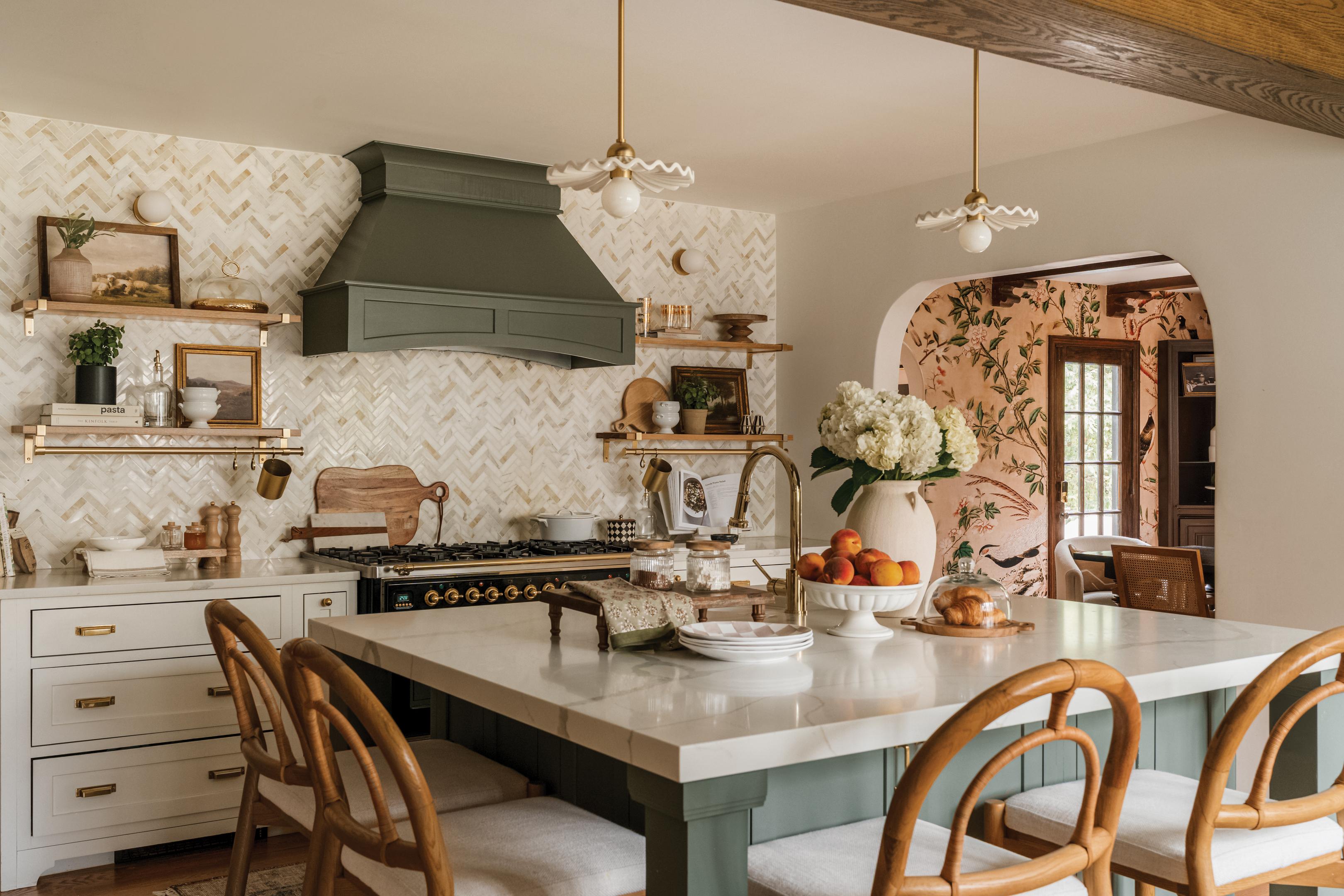Farmhouse kitchen with herringbone marble backsplash, sage green range hood, brass hardware, ruffled kitchen lights, and marble island with rattan bar stools.