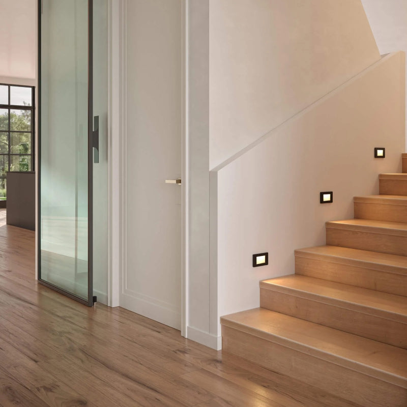 Modern staircase with warm recessed step lights installed along the wall, illuminating wooden stairs in a contemporary hallway.