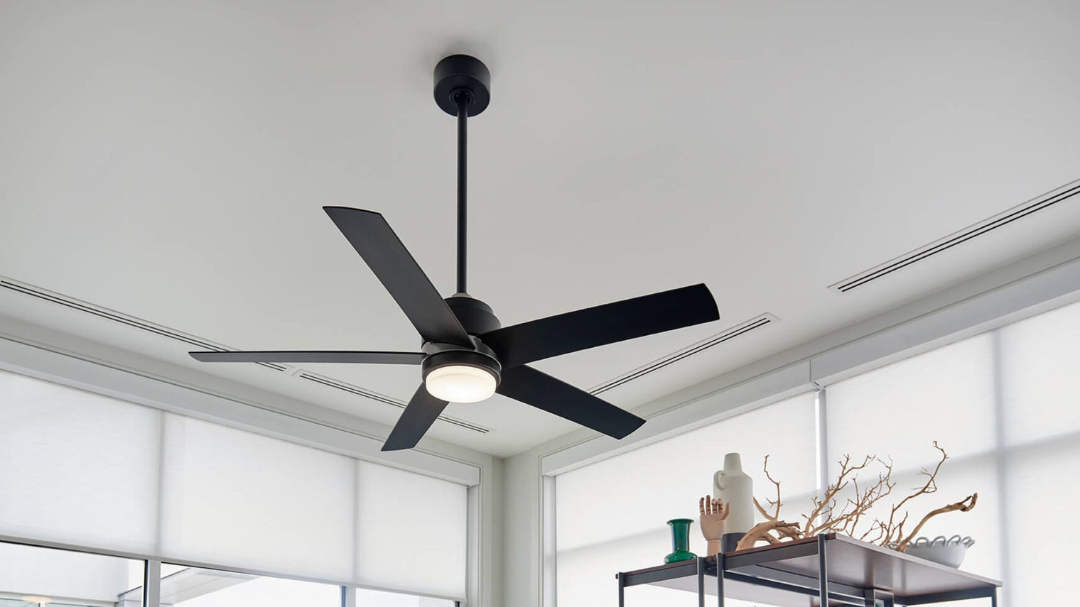 Matte black 5-blade ceiling fan with integrated LED light kit in bright modern living room with roller blinds