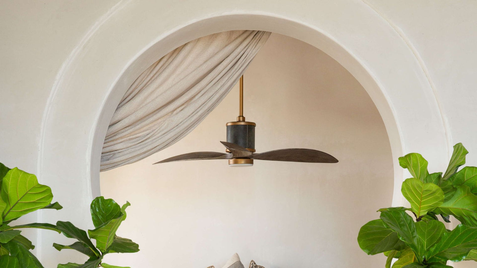 Two-blade dark wood and aged brass ceiling fan suspended in white plaster arched alcove with sheer draping and fiddle leaf fig plants