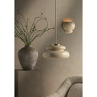 Styled room scene with cream disc pendant light on brass rod and ceramic bubble wall sconce, beside a vase with branches