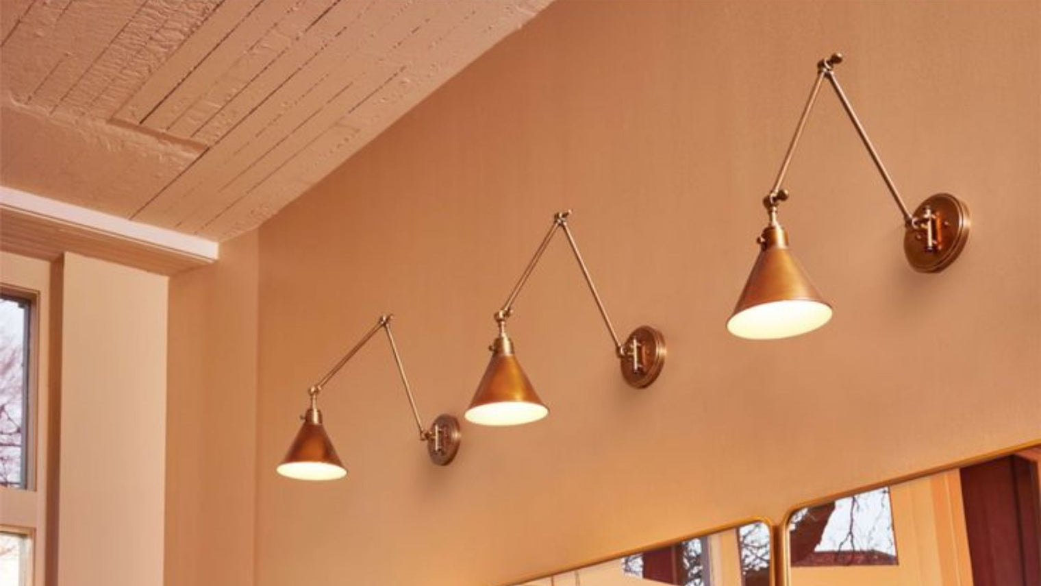 Three antique brass articulating swing arm wall sconces with cone shades at varying heights on warm terracotta wall with wood plank ceiling