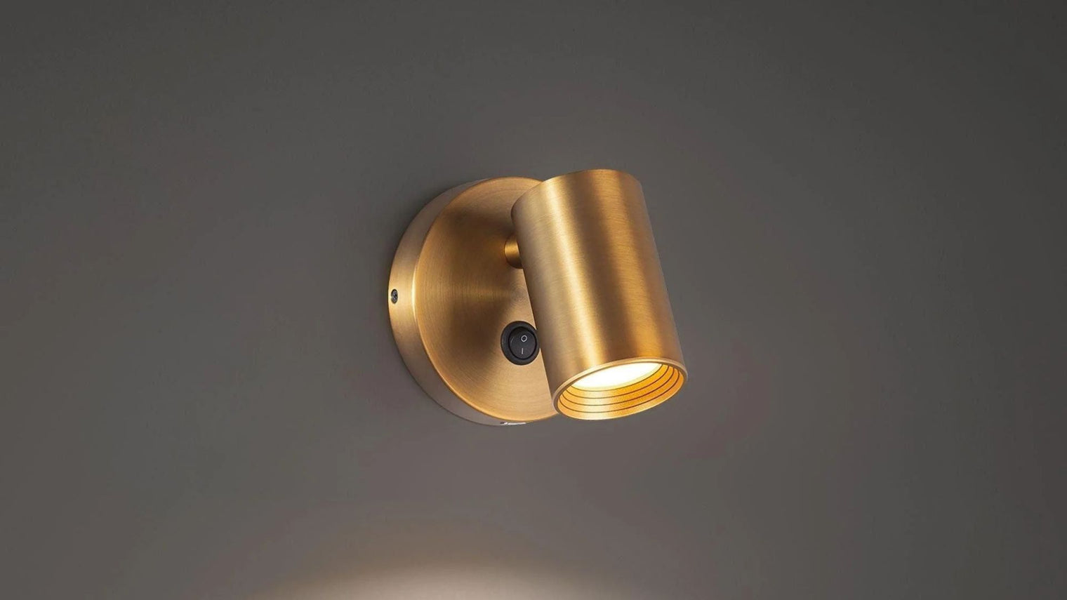 Brushed brass adjustable single cylinder spot wall sconce with toggle switch on grey wall
