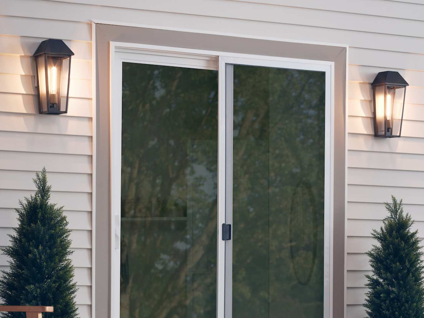 Pair of matte black traditional outdoor lantern wall sconces with exposed Edison filament bulbs flanking glass sliding patio door on cream clapboard exterior