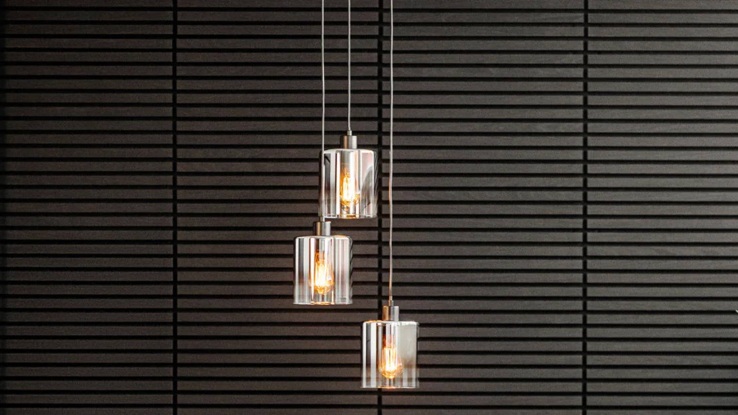 Trio of staggered brushed nickel pendants with ribbed clear glass cylinder shades and exposed Edison filament bulbs against dark slatted wall