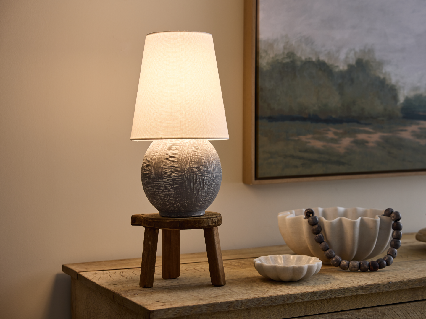 Textured grey ceramic globe table lamp on rustic three-legged wood stool base with white linen shade on reclaimed wood sideboard